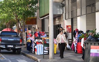 Mujer con chaqueta clara y pantalones oscuros lleva una bolsa en la mano, rodeada de expositores de camisetas en un ambiente urbano.