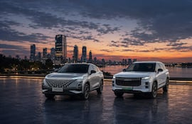 Dos SUV, uno gris y otro blanco, sobre una plataforma mirando la ciudad iluminada al atardecer.