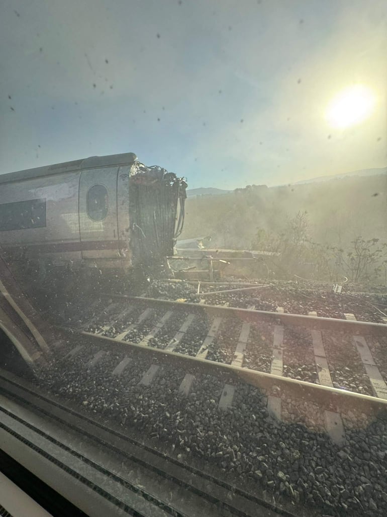 Tren accidentado con daños visibles en el vagón cinco, rodeado de escombros y vegetación, en un día soleado.