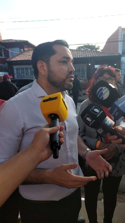 Luis Bello viste camisa blanca, conversa con periodistas que le apuntan micrófonos.