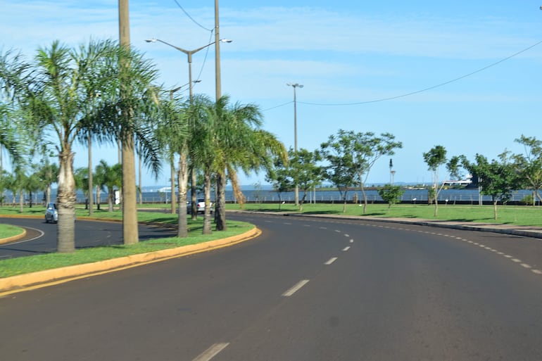 Carretera curvada flanqueada por palmeras y áreas verdes, sin personas, cielo azul claro y tráfico vehicular visible.