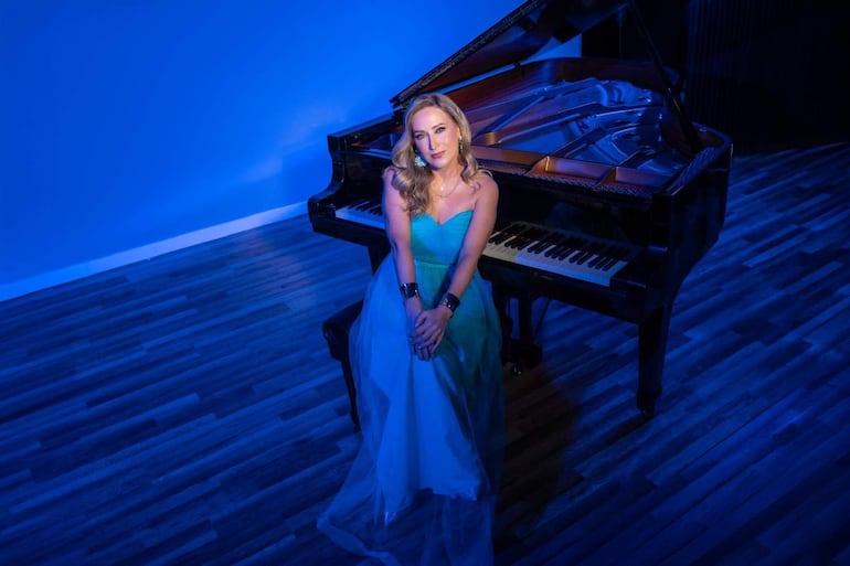 Mujer con vestido largo en gradiente de verde a azul, con cabello suelto, cruzando brazos junto a piano de cola iluminado.