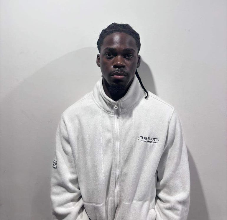 Joven Jabari Stephen Brown en chaqueta blanca, con cabello rizado y trenzas, mira serio hacia la cámara en un fondo blanco.