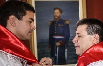 Santiago Peña y Horacio Cartes en camisas blancas con banda roja, ajustando distinción en ambiente institucional y formal.