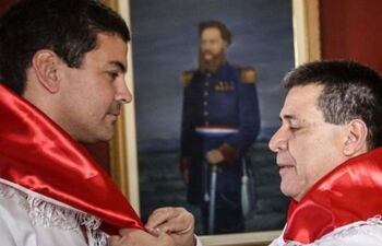Santiago Peña y Horacio Cartes en camisas blancas con banda roja, ajustando distinción en ambiente institucional y formal.