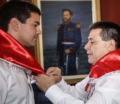 Santiago Peña y Horacio Cartes en camisas blancas con banda roja, ajustando distinción en ambiente institucional y formal.