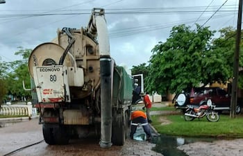 Dos trabajadores con chalecos de seguridad anaranjados, limpiando una zanja con agua estancada en un entorno residencial.