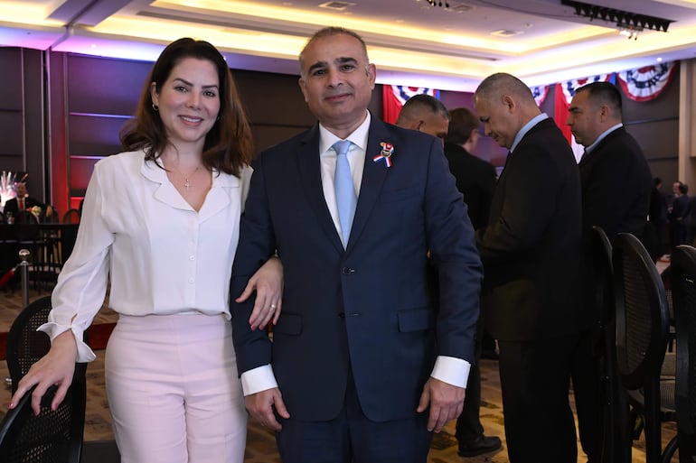 Pareja sonriente en evento formal; hombre en traje oscuro con corbata celeste y mujer en blusa blanca y pantalones claros.