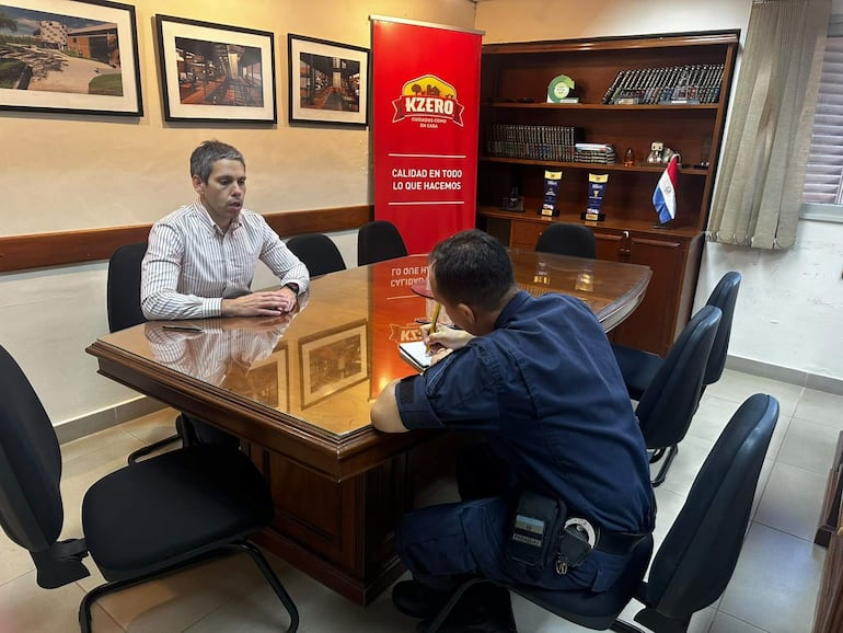 Hombre con camisa de rayas y persona en uniforme toman notas en oficina decorada con fotografías y banner de KZERO.