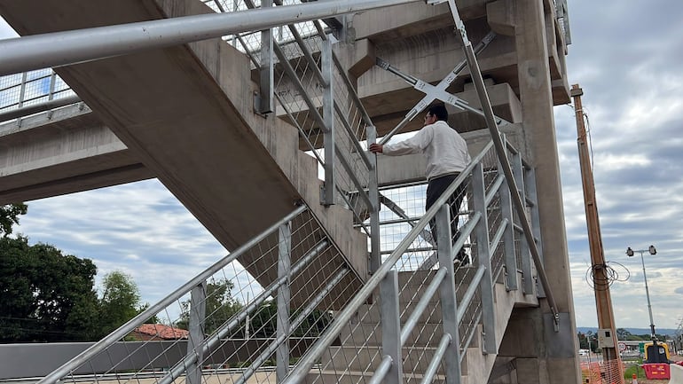 Hombre con camisa clara y pantalones oscuros subiendo escaleras metálicas en un entorno de construcción, cielo nublado detrás.