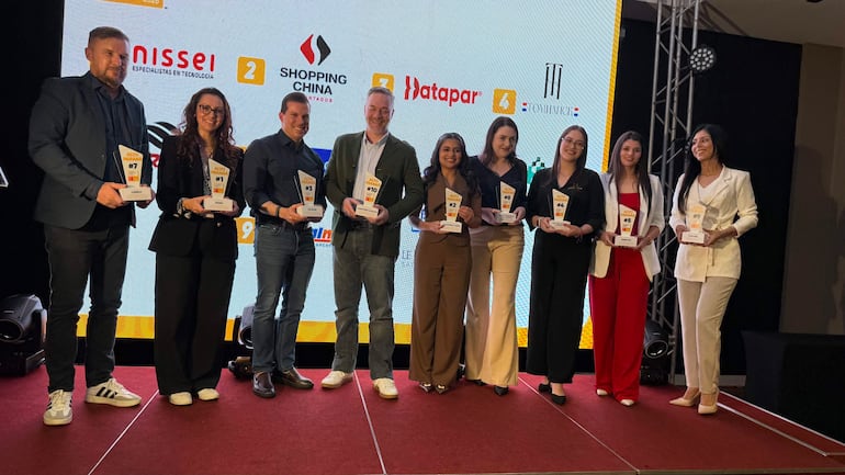 Ocho personas sostienen trofeos en un escenario iluminado; tres hombres y cinco mujeres lucen vestidos diversos en un ambiente formal.