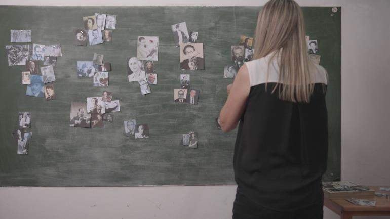Mujer de cabello largo y rubio, de espaldas, con blusa negra, observa un tablero verde lleno de fotografías dispersas en un entorno interior.