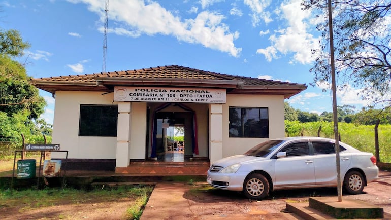 Comisaría N° 109 en entorno rural, edificio de una planta con automóvil gris claro estacionado y contenedor de reciclaje visible.