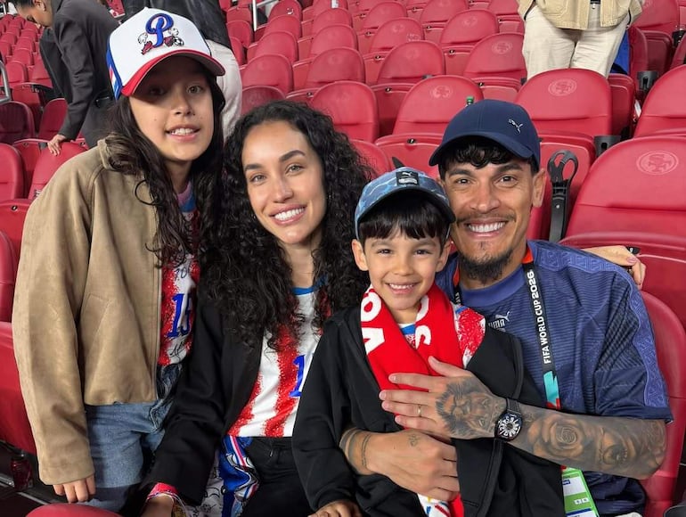Familia sonriente en estadio, hombre con tatuajes sostiene a un niño, ambos con bufandas. Niñas y mujer vestidas con camisetas de evento.
