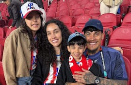 Familia sonriente en estadio, hombre con tatuajes sostiene a un niño, ambos con bufandas. Niñas y mujer vestidas con camisetas de evento.