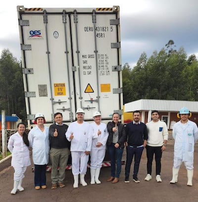 Grupo de ocho trabajadores, vestidos con ropa de trabajo, posan frente a un contenedor con gestos de alegría en un ambiente industrial.
