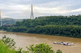 Bote rojo y blanco navega por río marrón con puente colgante al fondo y vegetación densa en orillas.