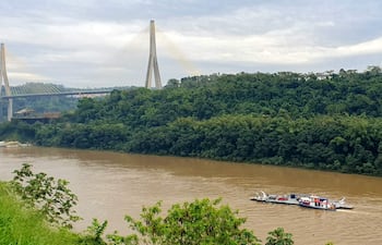 Bote rojo y blanco navega por río marrón con puente colgante al fondo y vegetación densa en orillas.