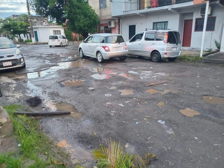 Calle en barrio Santa María con coches estacionados, pavimento deteriorado y charcos de agua, sin personas presentes.