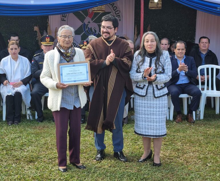 Isabel Báez Corvalán, anciana vestida elegantemente, sostiene un certificado mientras es aplaudida por Sergio Ríos y Elvina González de Díaz.
