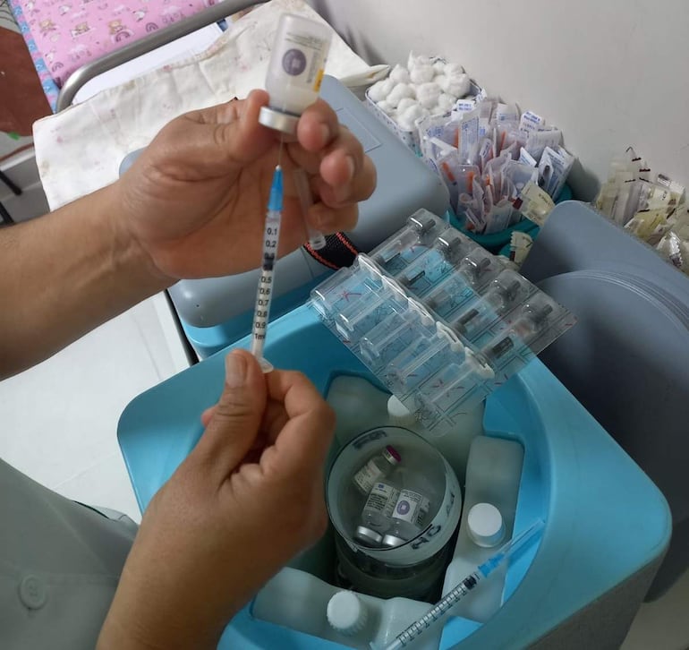 Persona concentrada en llenar jeringa con vacuna, en ambiente médico, rodeada de viales y jeringas.