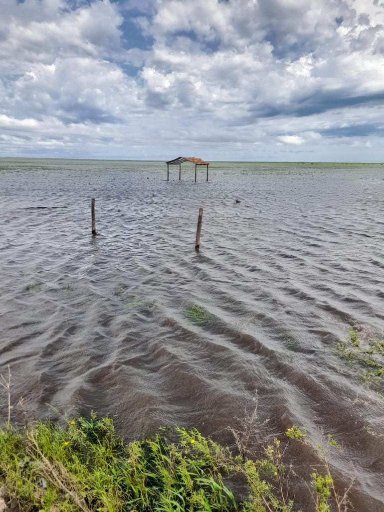 Paisaje inundado en Asentamiento Guayaki, con cielo nublado y una estructura de techo desgastada entre el agua.