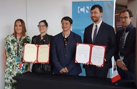 Christian Gayoso y Adriana Ortiz Semidei firman documentos, rodeados por otros adultos en ambiente formal con banner institucional.