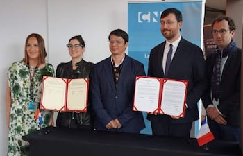 Christian Gayoso y Adriana Ortiz Semidei firman documentos, rodeados por otros adultos en ambiente formal con banner institucional.
