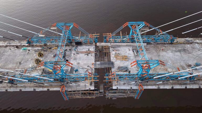 Vista aérea de un puente en construcción, con arcos y cables en azul y naranja, rodeado de escombros y herramientas.