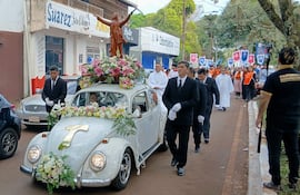 Dos hombres en trajes oscuros, uno conduciendo un Volkswagen adornado con flores, mientras siguen tunicas de colores en el desfile.