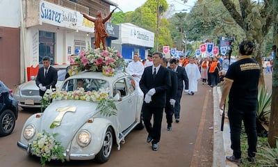 Dos hombres en trajes oscuros, uno conduciendo un Volkswagen adornado con flores, mientras siguen tunicas de colores en el desfile.