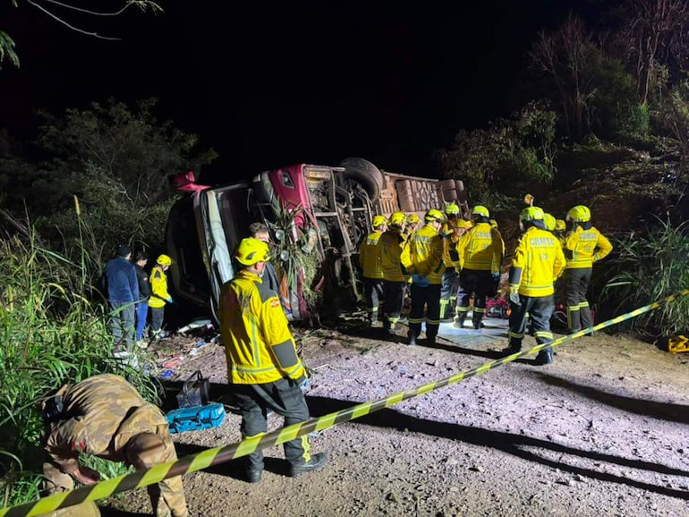 Rescatistas en uniformes amarillos rodean un bus volcado, asistiendo a una víctima en la carretera rodeada de vegetación.
