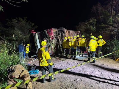Rescatistas en uniformes amarillos rodean un bus volcado, asistiendo a una víctima en la carretera rodeada de vegetación.
