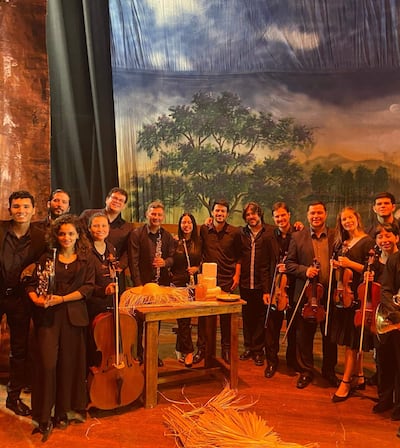 Un grupo de 15 músicos en negro, con violines y chelo, posan en un escenario con telón pintado de paisaje.
