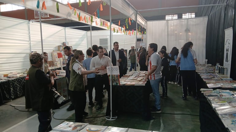 Mujer con chaleco verde interactúa con hombre de camisa blanca en Feria del Libro, rodeados de mesas con libros y decoraciones coloridas.