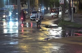 Motociclista con camiseta número 10 en calle mojada, rodeado de vehículos bajo luces de farolas en ambiente urbano.