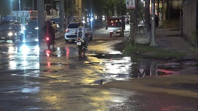 Motociclista con camiseta número 10 en calle mojada, rodeado de vehículos bajo luces de farolas en ambiente urbano.