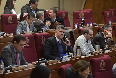 José Domingo Adorno Mazacotte sostiene un teléfono en medio de otros hombres en una sala de sesiones con escritorios de madera oscura.