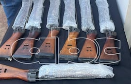 Hombre con chaleco táctico inspecciona fusiles y otras armas sobre una mesa en un entorno de presentación de evidencias.