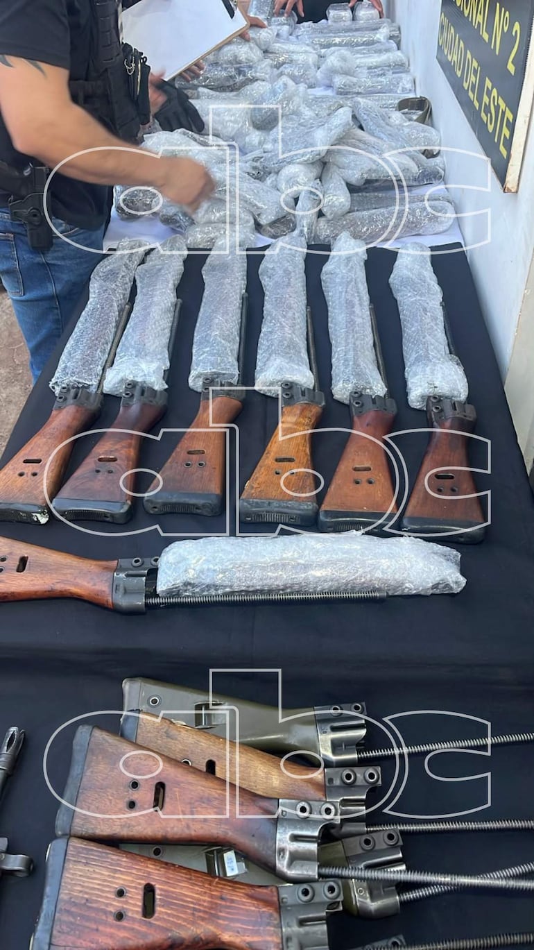 Hombre con chaleco táctico inspecciona fusiles y otras armas sobre una mesa en un entorno de presentación de evidencias.