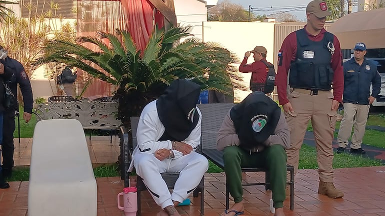 Dos hombres encapuchados sentados con manos sobre las piernas, mientras dos policías armados supervisan la situación en un entorno al aire libre.