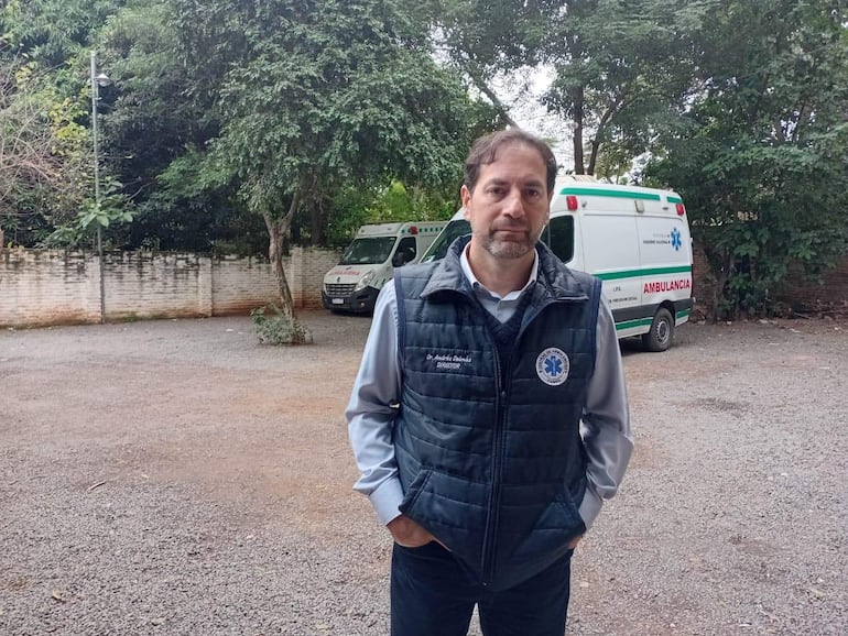 Hombre de pie en chaleco oscuro y camisa, rodeado de vegetación, con dos ambulancias al fondo.