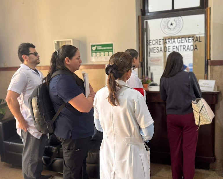 Cinco personas, dos con batas blancas, dialogan en la ventanilla de Secretaría General, con ambiente de oficina y luz natural.