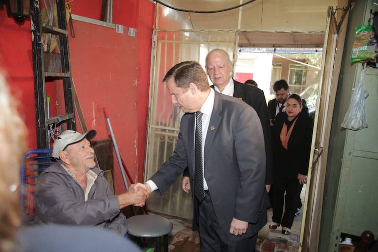 Dos hombres en trajes oscuros, uno apretando manos con un hombre mayor. Ambiente informal con paredes rojas.