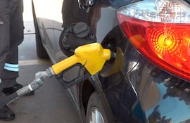 Hombre con uniforme gris y franjas reflectantes sostiene manguera amarilla en una gasolinera, con un auto negro como fondo.