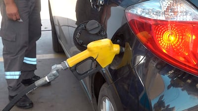 Hombre con uniforme gris y franjas reflectantes sostiene manguera amarilla en una gasolinera, con un auto negro como fondo.