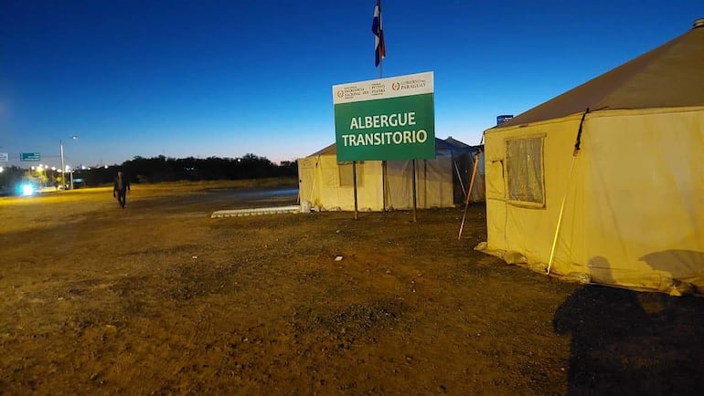 Albergue transitorio con cartel verde y blanco, en un entorno de tierra y arena rodeado de vegetación baja.