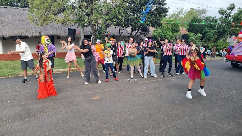 Niña disfrazada y clown bailan en un ambiente festivo rodeados de jóvenes con camisetas a rayas rojas y blancas.