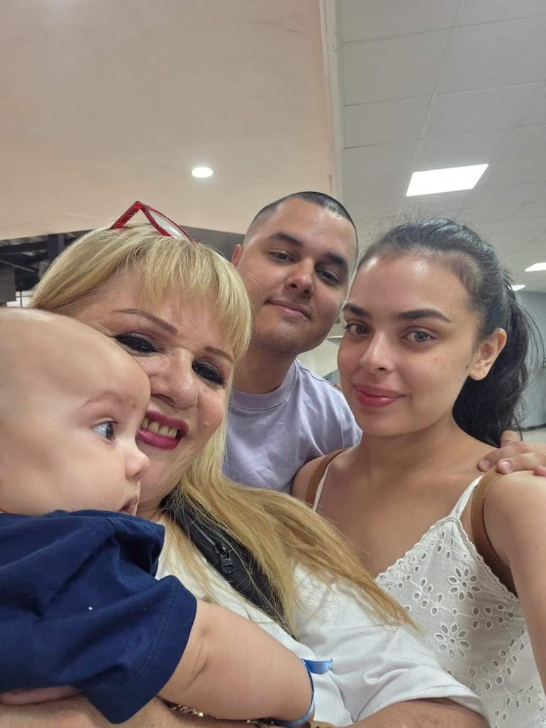 Mujer rubia sonriente sostiene un bebé; hombre de cabello oscuro sonriendo a su lado; joven con bolsa mira a la cámara.