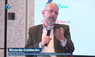 Ricardo Calderón sentado, con micrófono en mano, vistiendo saco oscuro y suéter blanco, hablando en un evento de periodismo.
